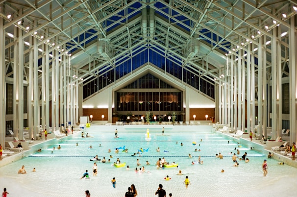 A spacious indoor pool area featuring a high, glass-paneled ceiling and well-lit surroundings. Numerous people of various ages are swimming, floating, or relaxing in the large swimming pool. The architectural design is modern and open, with tall supporting beams and a symmetrical layout. There are pool chairs lining the sides and a few decorations, such as potted plants, near the back wall.