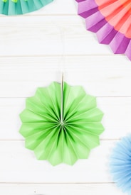 A variety of colorful, folded paper fans arranged around a central green fan, all placed on a light wooden surface. The fans are circular with pleated patterns in pastel colors including green, pink, purple, and light blue.