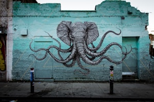 A mural on a brick wall depicts a surreal combination of an elephant's head with octopus tentacles extending outward. The artwork is painted on a weathered turquoise brick wall in an urban setting, with two bollards in front and some graffiti on nearby surfaces.