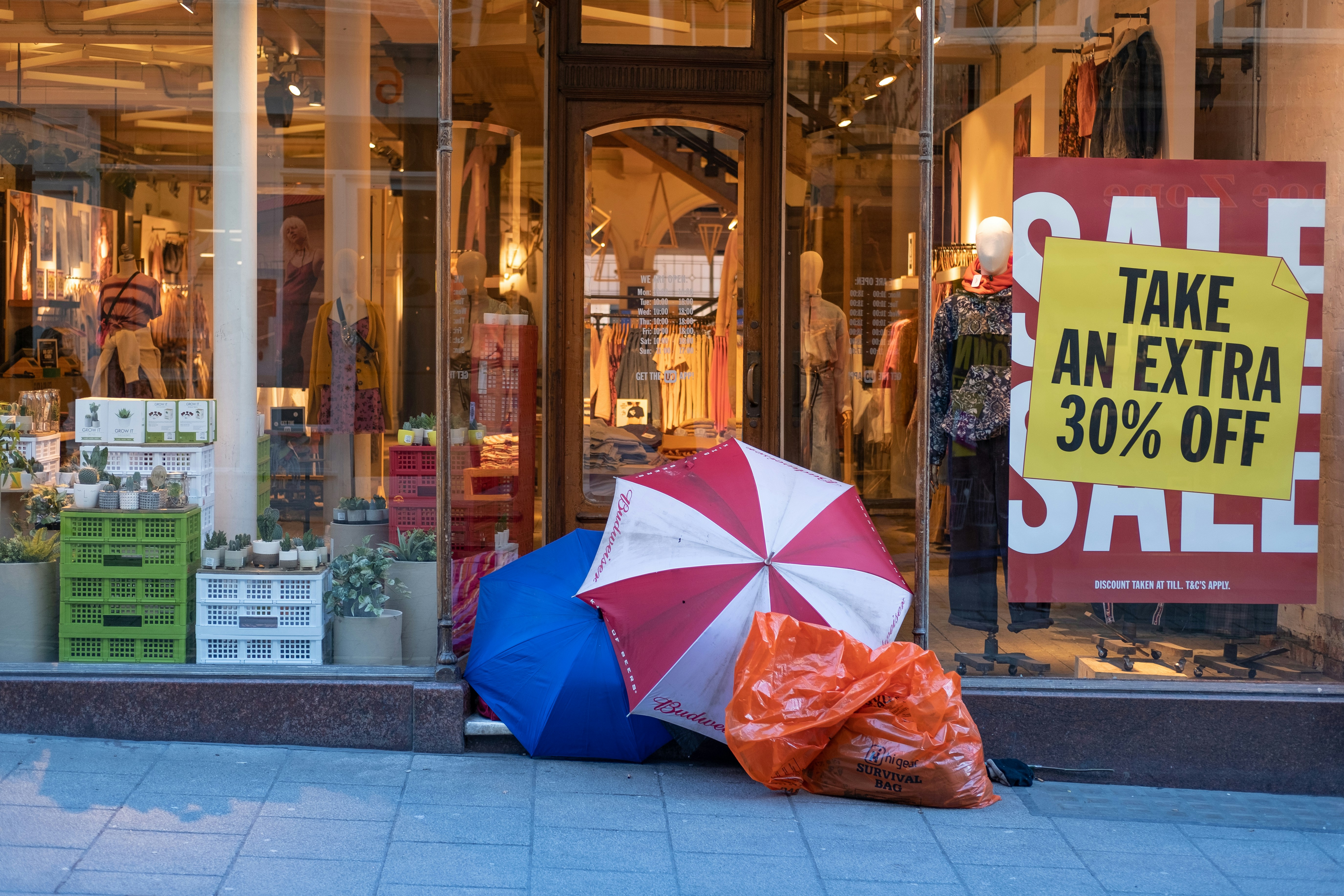 A homeless man takes shelter under opened umbrellas in the front door of a retail shop, while the shop next to him offers significant sales discounts. A juxtaposition - excess and having nothing. The lighting in the shot (the warm hues of the inside of the shop) compound the feeling of coldness on the outside.