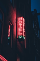 A neon sign on the side of a building displays the word 'HOTEL' in bright pink and green lights. The number '83' is also lit up in neon green. The building appears to be a dark brick structure, slightly illuminated by the glow of the sign. The sky is dark, suggesting it is nighttime.