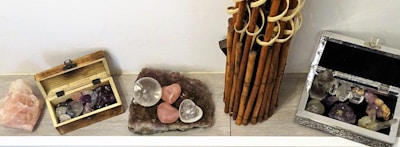 A display of various crystals and stones arranged on a flat surface. There are two wooden boxes, one open showing an assortment of small polished stones. There's also a cluster of rose quartz hearts and spheres on a stone base. An arrangement of bamboo stalks stands upright alongside these items.