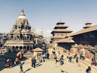 A bustling historical square featuring intricately carved stone temples with multi-tiered roofs and traditional Newari architecture. People are walking and gathering around the open space, creating a lively atmosphere. The sky is clear, providing a bright backdrop to the scene.