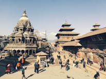 A bustling historical square featuring intricately carved stone temples with multi-tiered roofs and traditional Newari architecture. People are walking and gathering around the open space, creating a lively atmosphere. The sky is clear, providing a bright backdrop to the scene.