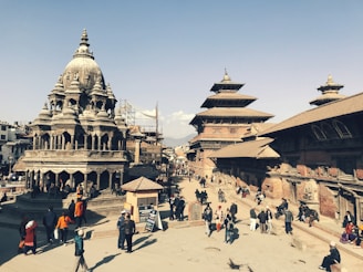A bustling historical square featuring intricately carved stone temples with multi-tiered roofs and traditional Newari architecture. People are walking and gathering around the open space, creating a lively atmosphere. The sky is clear, providing a bright backdrop to the scene.