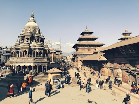 A bustling historical square featuring intricately carved stone temples with multi-tiered roofs and traditional Newari architecture. People are walking and gathering around the open space, creating a lively atmosphere. The sky is clear, providing a bright backdrop to the scene.