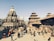 A bustling historical square featuring intricately carved stone temples with multi-tiered roofs and traditional Newari architecture. People are walking and gathering around the open space, creating a lively atmosphere. The sky is clear, providing a bright backdrop to the scene.