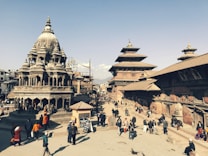 A bustling historical square featuring intricately carved stone temples with multi-tiered roofs and traditional Newari architecture. People are walking and gathering around the open space, creating a lively atmosphere. The sky is clear, providing a bright backdrop to the scene.