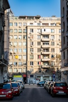 A street scene in an urban environment with a large apartment building in the background. The building features multiple balconies and numerous air conditioning units. On the ground level, there are small businesses, including a florist shop. Several parked cars line the street, and a few people are visible in the distance.