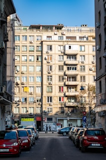 A street scene in an urban environment with a large apartment building in the background. The building features multiple balconies and numerous air conditioning units. On the ground level, there are small businesses, including a florist shop. Several parked cars line the street, and a few people are visible in the distance.