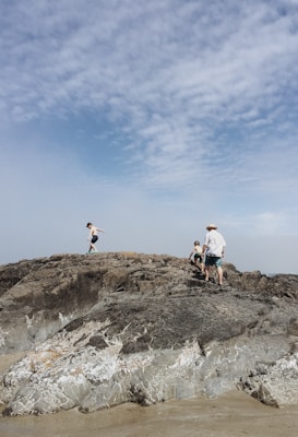 Children and an adult are walking on large, rugged rocks by the seaside. The sky is partly cloudy, creating a serene and relaxed atmosphere.