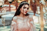 A woman dressed in an elegant, floral lace gown stands in a decorative outdoor setting, adorned with a delicate headpiece made of flowers. The background includes blurred natural elements and strands of flowers and greenery, creating a soft and romantic atmosphere.