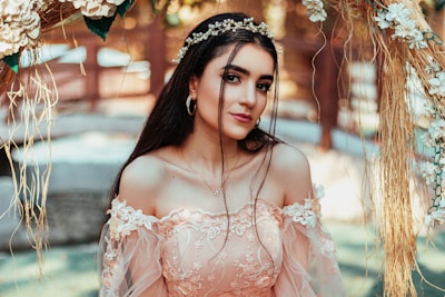A woman dressed in an elegant, floral lace gown stands in a decorative outdoor setting, adorned with a delicate headpiece made of flowers. The background includes blurred natural elements and strands of flowers and greenery, creating a soft and romantic atmosphere.