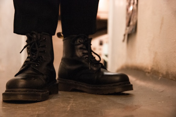A pair of black leather boots with laces is worn by a person standing indoors. The boots have a rugged appearance, and the view is from a low angle focusing on the footwear.
