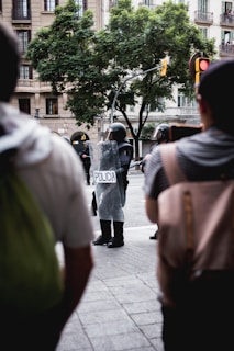 A group of police officers in riot gear is standing in the street, with shields labeled 'POLICIA.' The scene takes place in an urban setting with a large tree and an apartment building in the background. Several people are observing from the foreground and the atmosphere suggests a public demonstration or gathering.
