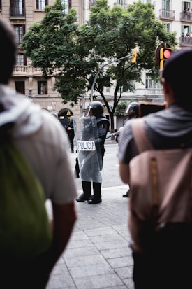 A group of police officers in riot gear is standing in the street, with shields labeled 'POLICIA.' The scene takes place in an urban setting with a large tree and an apartment building in the background. Several people are observing from the foreground and the atmosphere suggests a public demonstration or gathering.
