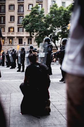 A group of police officers in riot gear holding shields labeled 'policia' form a line on a city street. In the foreground, a person kneels on the sidewalk, facing the police. The backdrop includes a multi-story building and lush green trees.