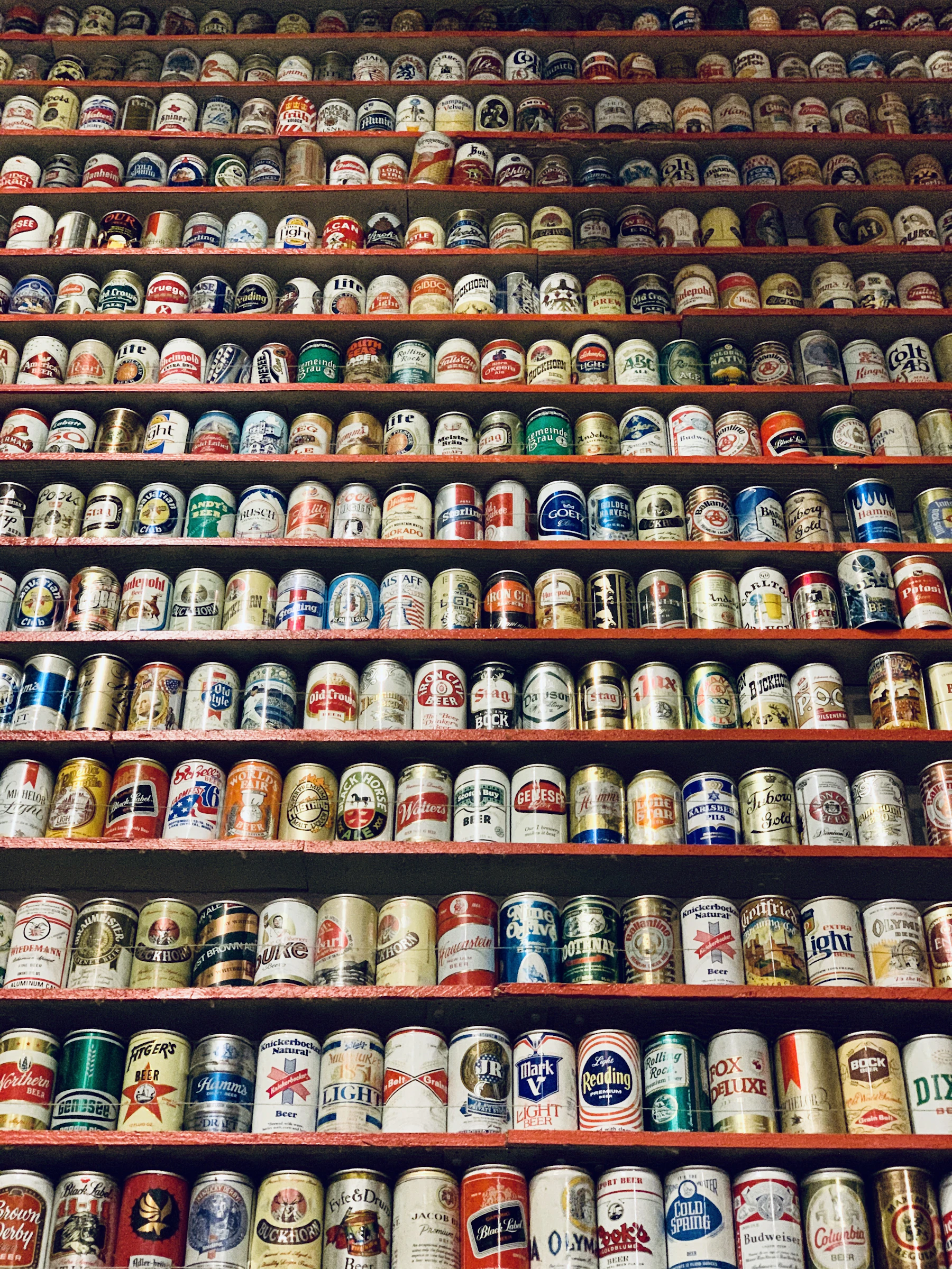 A wall of vintage beer cans standing in rows on red shelves from floor to ceiling