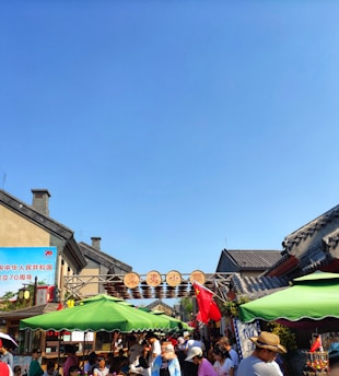 A busy street market with many people gathering under colorful umbrellas and banners. Traditional Chinese architecture frames the scene, and there are signs in both Chinese characters and a red flag. Bright sunlight casts shadows, and the sky is a clear blue, creating a lively and vibrant atmosphere.