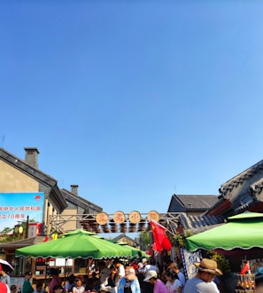 A busy street market with many people gathering under colorful umbrellas and banners. Traditional Chinese architecture frames the scene, and there are signs in both Chinese characters and a red flag. Bright sunlight casts shadows, and the sky is a clear blue, creating a lively and vibrant atmosphere.