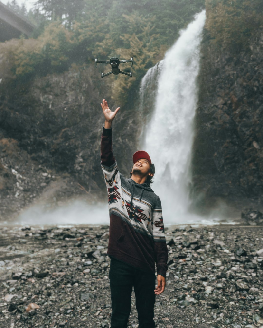 man reaching flying quadcopter near water fall