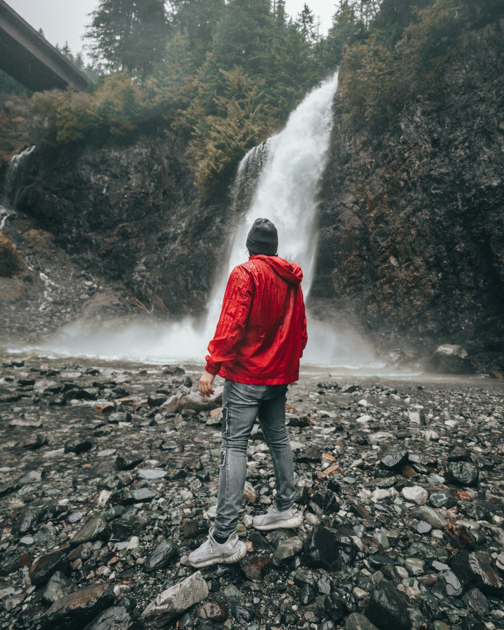 man in red jacket facing waterfall during daytime