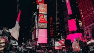 A vibrant urban scene featuring illuminated billboards and advertisements in a bustling city area. The buildings are adorned with large electronic displays showcasing various brands and products. Pedestrians and vehicles are visible on the street, adding to the lively atmosphere.