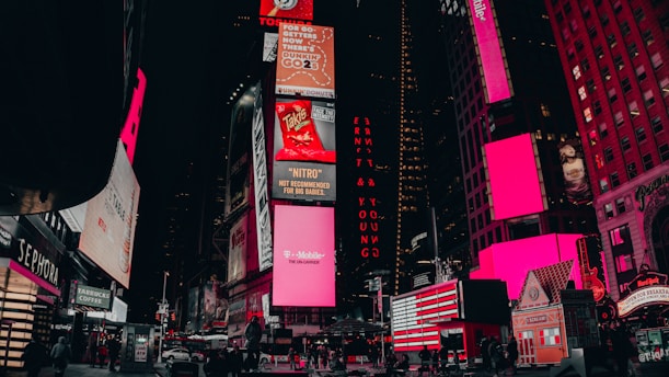 A vibrant urban scene featuring illuminated billboards and advertisements in a bustling city area. The buildings are adorned with large electronic displays showcasing various brands and products. Pedestrians and vehicles are visible on the street, adding to the lively atmosphere.