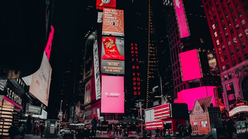 A vibrant urban scene featuring illuminated billboards and advertisements in a bustling city area. The buildings are adorned with large electronic displays showcasing various brands and products. Pedestrians and vehicles are visible on the street, adding to the lively atmosphere.