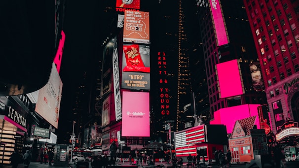 A vibrant urban scene featuring illuminated billboards and advertisements in a bustling city area. The buildings are adorned with large electronic displays showcasing various brands and products. Pedestrians and vehicles are visible on the street, adding to the lively atmosphere.