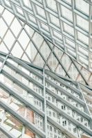 A structural glass facade with a grid-like pattern of metal frames intersecting at angles, creating a geometric design. The transparent glass allows views of the buildings outside, which are partially visible through the grid, adding depth and dimension to the composition.