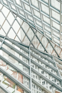 A structural glass facade with a grid-like pattern of metal frames intersecting at angles, creating a geometric design. The transparent glass allows views of the buildings outside, which are partially visible through the grid, adding depth and dimension to the composition.