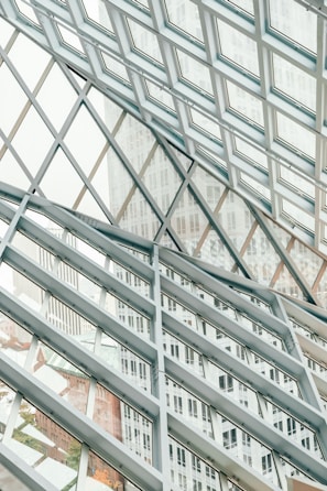 A structural glass facade with a grid-like pattern of metal frames intersecting at angles, creating a geometric design. The transparent glass allows views of the buildings outside, which are partially visible through the grid, adding depth and dimension to the composition.