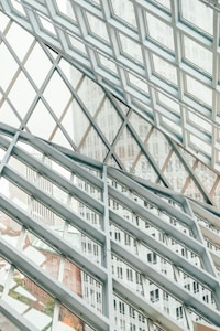 A structural glass facade with a grid-like pattern of metal frames intersecting at angles, creating a geometric design. The transparent glass allows views of the buildings outside, which are partially visible through the grid, adding depth and dimension to the composition.