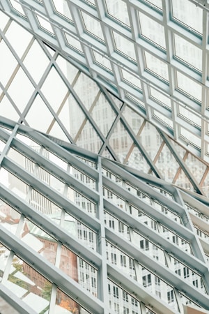 A structural glass facade with a grid-like pattern of metal frames intersecting at angles, creating a geometric design. The transparent glass allows views of the buildings outside, which are partially visible through the grid, adding depth and dimension to the composition.