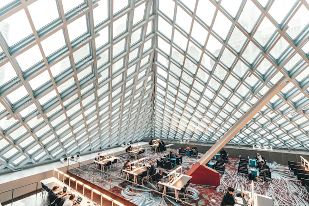 A modern library space featuring a large, geometric glass ceiling that floods the area with natural light. People are seated at desks, working on laptops or reading, creating a focused and studious atmosphere. The floor is carpeted with a colorful, abstract pattern, and the architecture is characterized by sleek lines and a spacious layout.