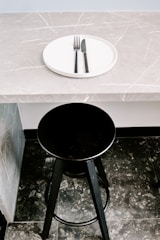 A black bar stool is positioned in front of a gray marble countertop with a subtle veined pattern. On the countertop rests a white circular plate with a black fork and knife neatly placed on it.