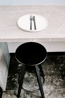 A black bar stool is positioned in front of a gray marble countertop with a subtle veined pattern. On the countertop rests a white circular plate with a black fork and knife neatly placed on it.