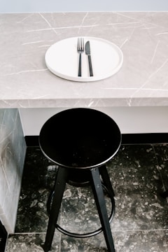 A black bar stool is positioned in front of a gray marble countertop with a subtle veined pattern. On the countertop rests a white circular plate with a black fork and knife neatly placed on it.