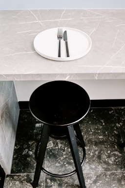 A black bar stool is positioned in front of a gray marble countertop with a subtle veined pattern. On the countertop rests a white circular plate with a black fork and knife neatly placed on it.