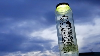 A transparent bottle with a black cap is prominently featured against a cloudy sky. The bottle has a playful cartoon monkey illustration and various inspirational words like 'SMILE', 'DREAM', 'LOVE', and 'CHILL' printed on it. The bottle appears to be partially filled with a liquid.