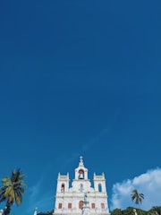 A white church building with ornate architecture stands prominently against a bright blue sky. The building features several spires, arched windows, and a statue at the front. Tall palm trees are situated on both sides of the building, adding a tropical feel to the scene.