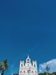 A white church building with ornate architecture stands prominently against a bright blue sky. The building features several spires, arched windows, and a statue at the front. Tall palm trees are situated on both sides of the building, adding a tropical feel to the scene.