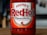A bottle of Frank's RedHot Original Cayenne Pepper Sauce, featuring a bold red label with white and red lettering. The label also includes a graphic of chili peppers and text stating 'The Perfect Blend of Flavour & Heat'. The bottle sits on a wooden surface with a blurred background.