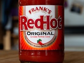 A bottle of Frank's RedHot Original Cayenne Pepper Sauce, featuring a bold red label with white and red lettering. The label also includes a graphic of chili peppers and text stating 'The Perfect Blend of Flavour & Heat'. The bottle sits on a wooden surface with a blurred background.