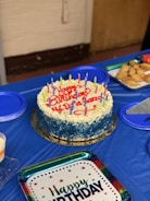 A round birthday cake with blue and white frosting is topped with colorful candles and the words 'Happy Birthday' written in red icing. It is placed on a blue tablecloth surrounded by plates, some filled with pastries. A 'Happy Birthday' plate is also visible.
