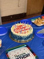 A round birthday cake with blue and white frosting is topped with colorful candles and the words 'Happy Birthday' written in red icing. It is placed on a blue tablecloth surrounded by plates, some filled with pastries. A 'Happy Birthday' plate is also visible.