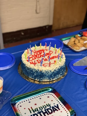 A round birthday cake with blue and white frosting is topped with colorful candles and the words 'Happy Birthday' written in red icing. It is placed on a blue tablecloth surrounded by plates, some filled with pastries. A 'Happy Birthday' plate is also visible.