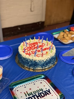 A round birthday cake with blue and white frosting is topped with colorful candles and the words 'Happy Birthday' written in red icing. It is placed on a blue tablecloth surrounded by plates, some filled with pastries. A 'Happy Birthday' plate is also visible.