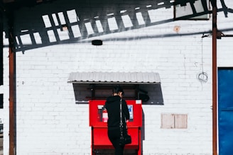 A person dressed in black is standing in front of a red machine mounted on a white brick wall. The machine has a small roof above it. Shadows of an overhead structure create patterns on the white wall.